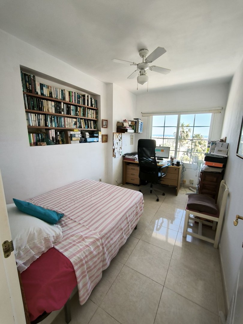 Bedroom with a bed, built-in bookshelves, a desk with a computer, and a large window overlooking palm trees and the sea.