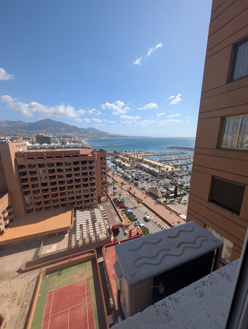 View from a balcony with a tennis court below and a scenic sea view with mountains in the distance.