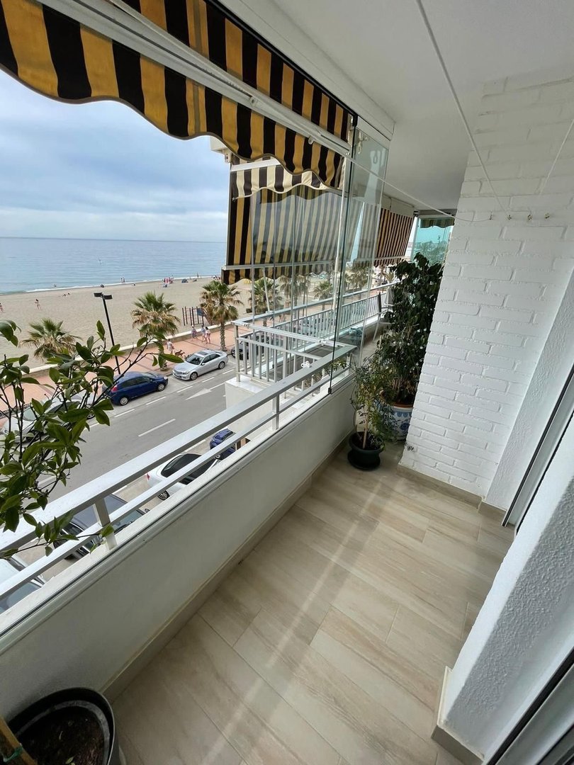 A spacious balcony with a sea view, featuring an awning, plants, and glass railings overlooking a beach and street.
