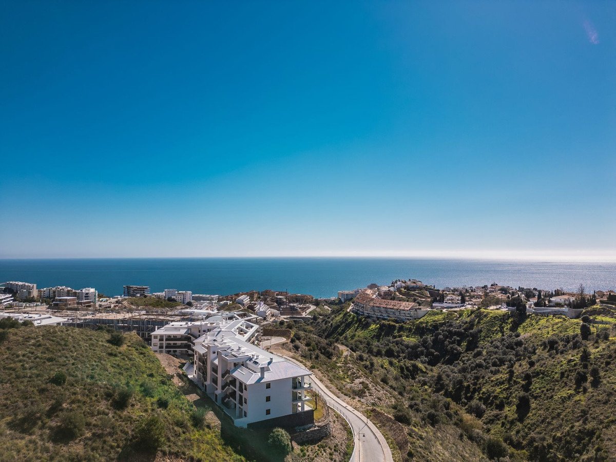 Modern apartment complex with balconies, overlooking the sea and a hillside town under a clear blue sky.