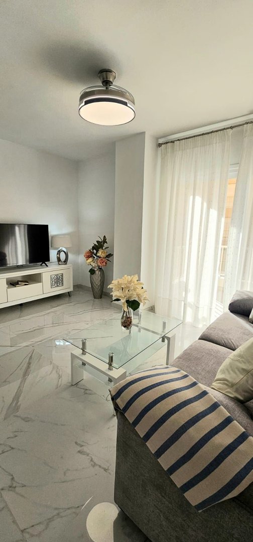 Modern living room with a grey sofa, glass coffee table, TV, and large window with sheer curtains.