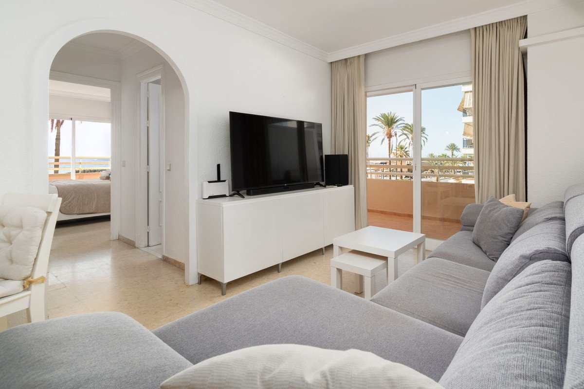 Modern living room with grey sofa, TV, and balcony overlooking palm trees and the sea.