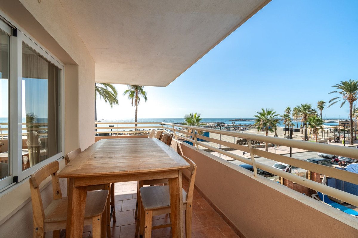 Wooden dining table and chairs on a balcony overlooking a beach with palm trees and the sea.