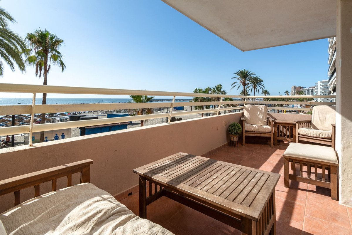 A spacious balcony with wooden furniture and tables, offering a clear view of the beach, palm trees, and the sea.