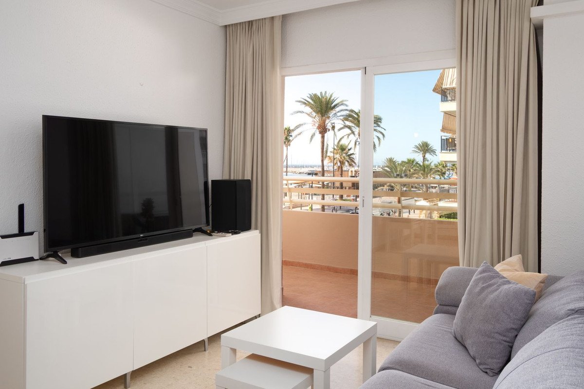 A living room with a large TV, sofa, and a sliding glass door opening to a balcony with palm trees and a sea view.