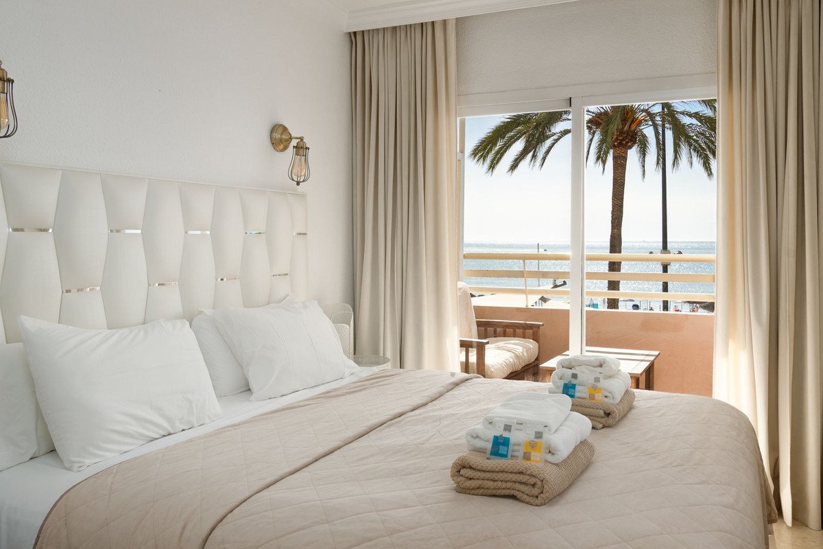 A hotel bedroom with a large bed and a balcony door offering an ocean view with a palm tree.