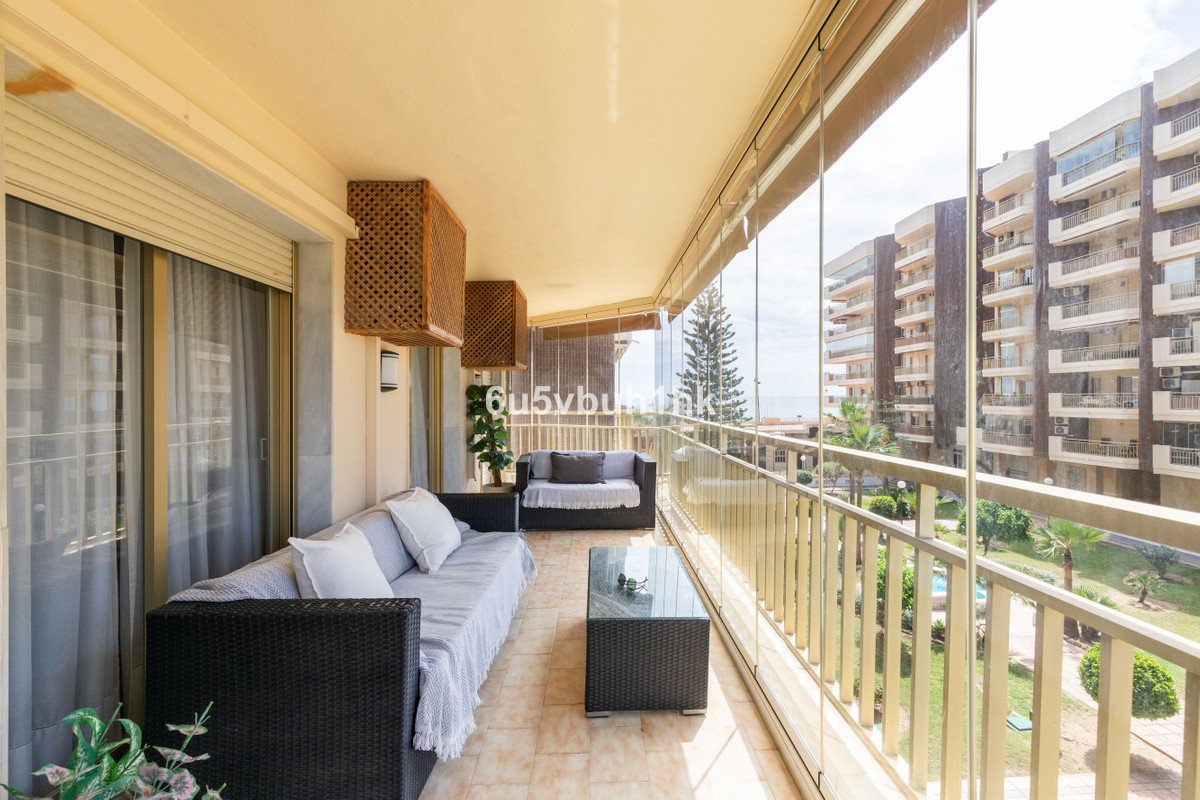 Enclosed balcony with sofas, coffee table, and view of buildings, trees, and the sea.