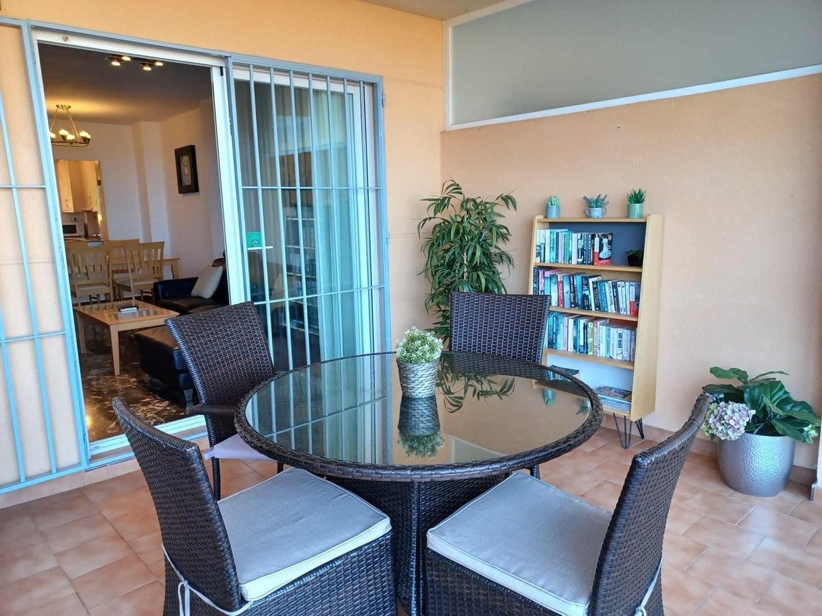 A round glass-top table with four rattan chairs on a balcony, featuring a bookshelf and plants.