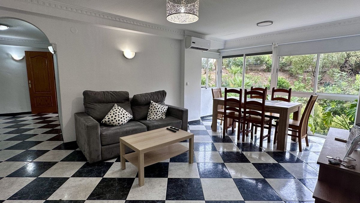 A spacious living and dining area with a sofa, coffee table, and a dining set, featuring a large window with a lush garden view.