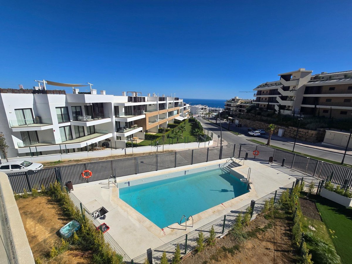 A bright blue swimming pool is visible with a modern apartment complex in the background and the sea in the distance.