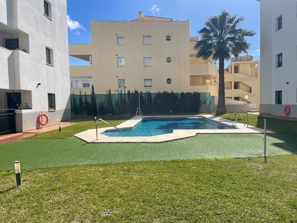 Outdoor view of a communal swimming pool surrounded by artificial grass, palm trees, and residential buildings under a blue sky.