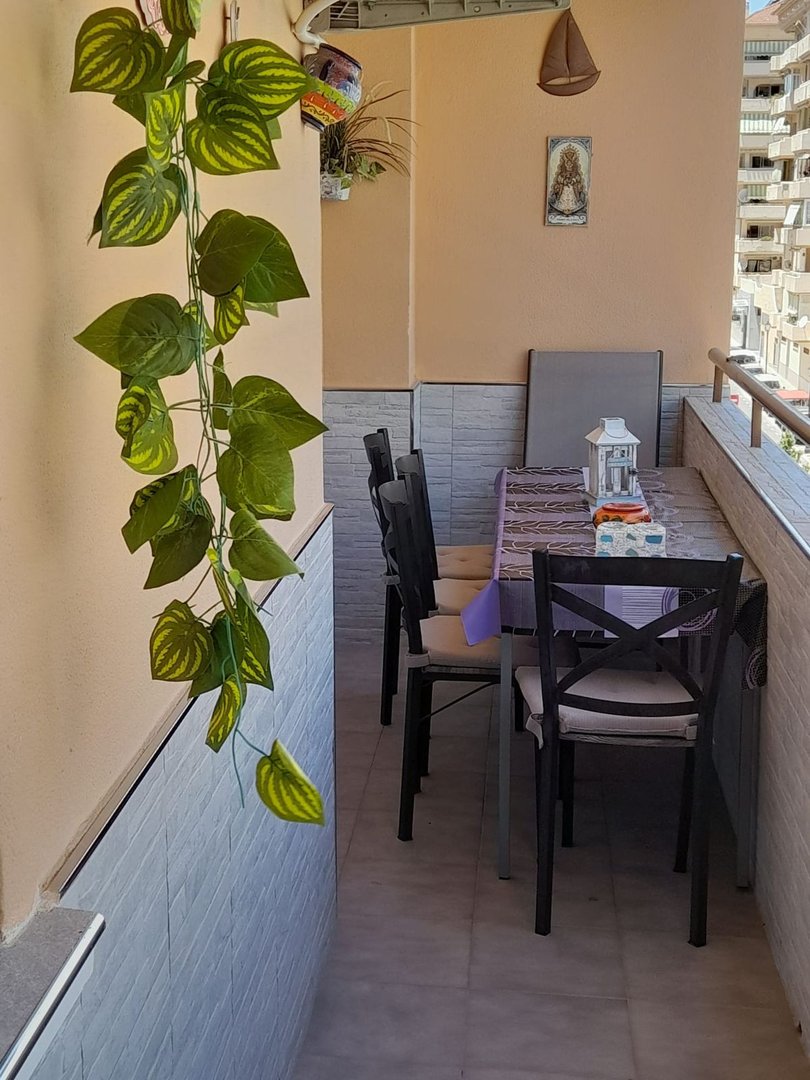 A narrow balcony with a table, chairs, and a hanging plant overlooking an urban cityscape.