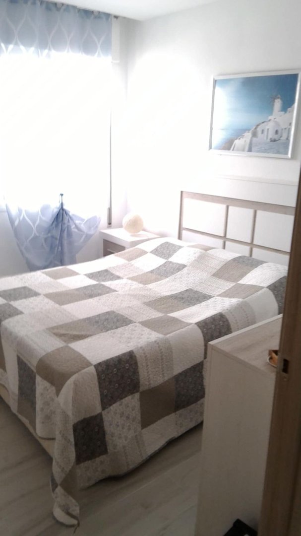 A bedroom with a quilted bedspread, a nightstand with a lamp, and a framed artwork of white buildings by the sea.