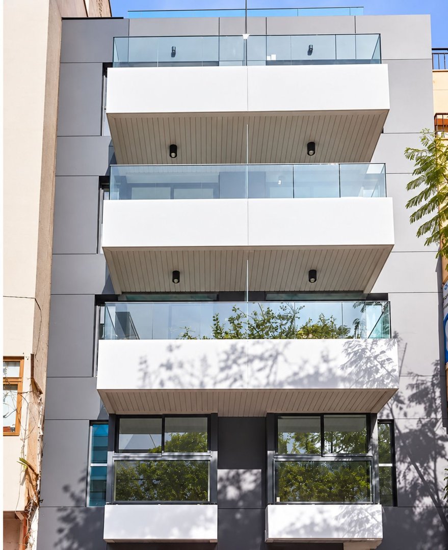 Modern apartment building facade with multiple balconies featuring glass railings and white overhangs, reflecting green trees.