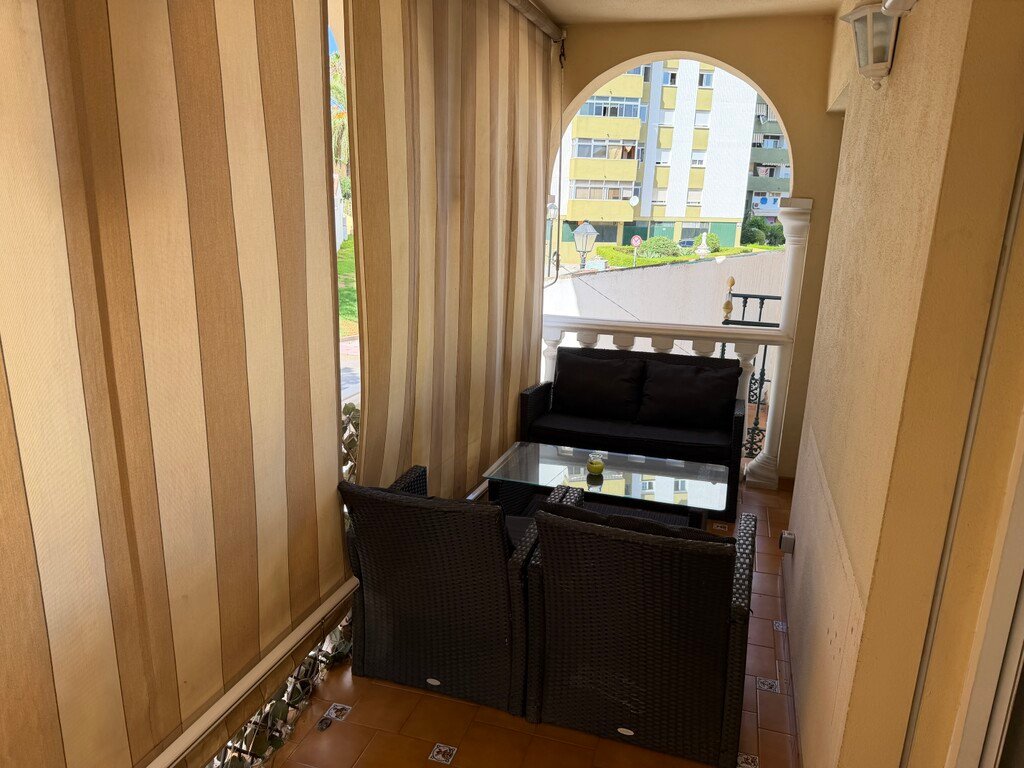Balcony with dark rattan furniture, glass coffee table, and striped awnings. Overlooks buildings and greenery.