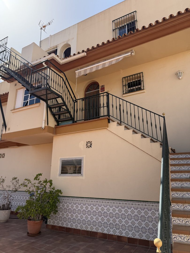 Exterior view of a building with stairs, balcony railings, potted plants, and patterned tiles.