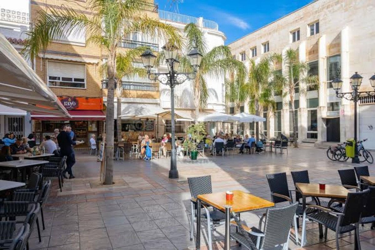 An outdoor cafe terrace with tables and chairs under umbrellas, palm trees, and people dining.