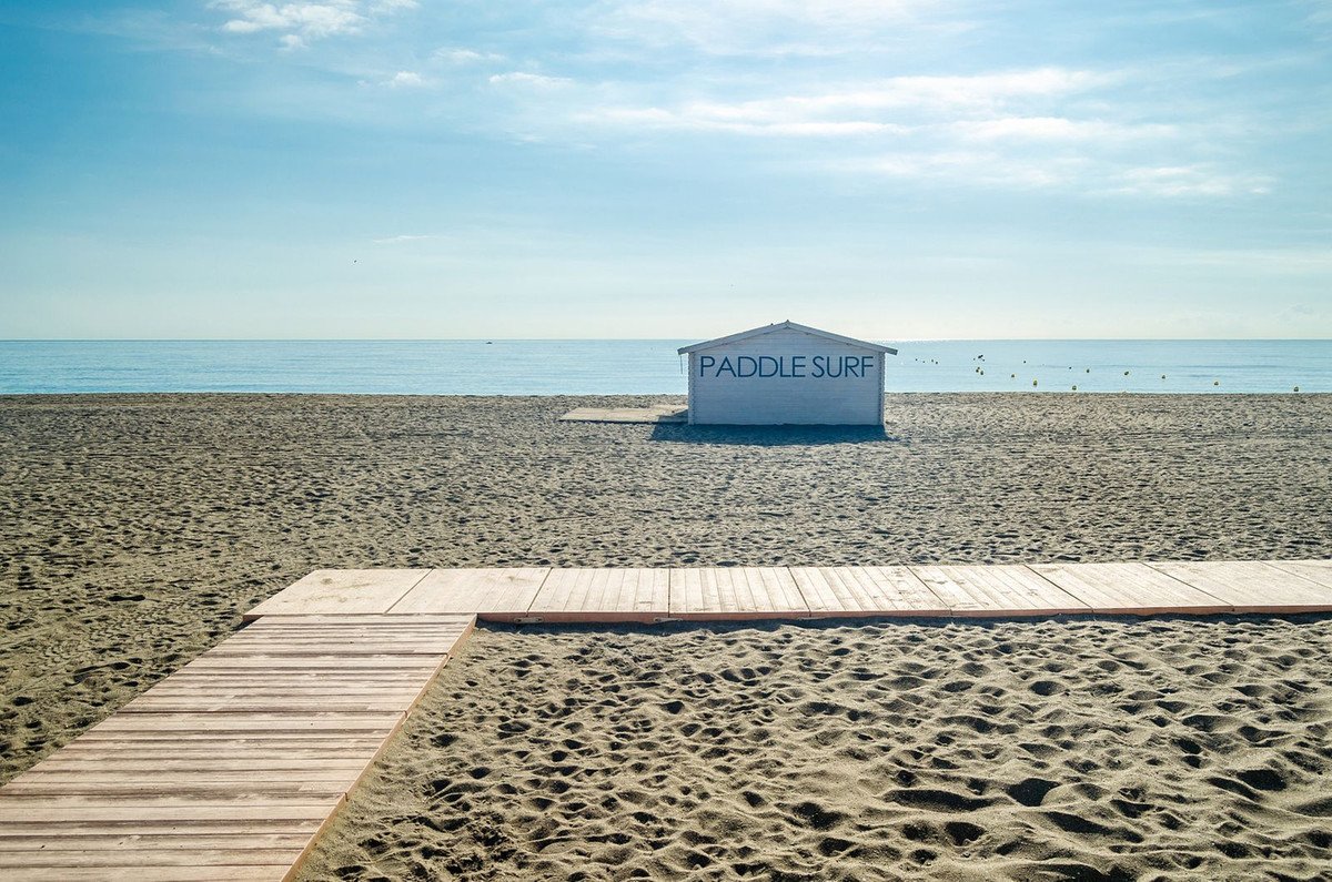 A white beach hut labeled 'PADDLE SURF' sits on a sandy beach with a wooden walkway leading towards the calm blue sea.