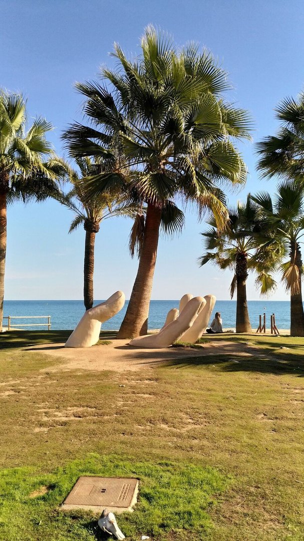 A large sculpture of a hand is situated on a grassy area overlooking the ocean, surrounded by palm trees.