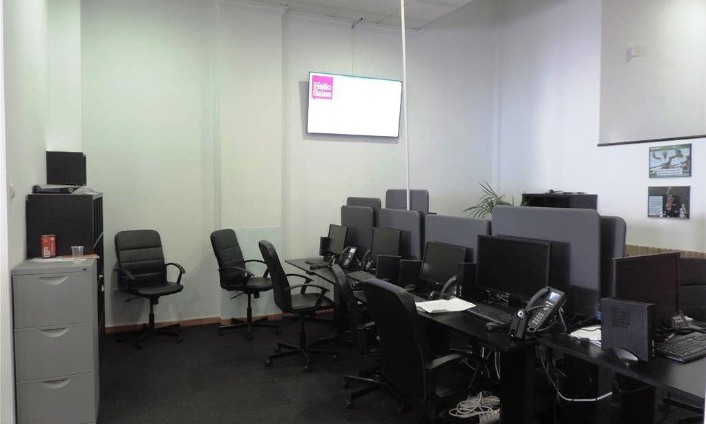 A modern office with multiple computer workstations, including monitors, keyboards, phones, and chairs, with a large screen displaying a logo on the wall.