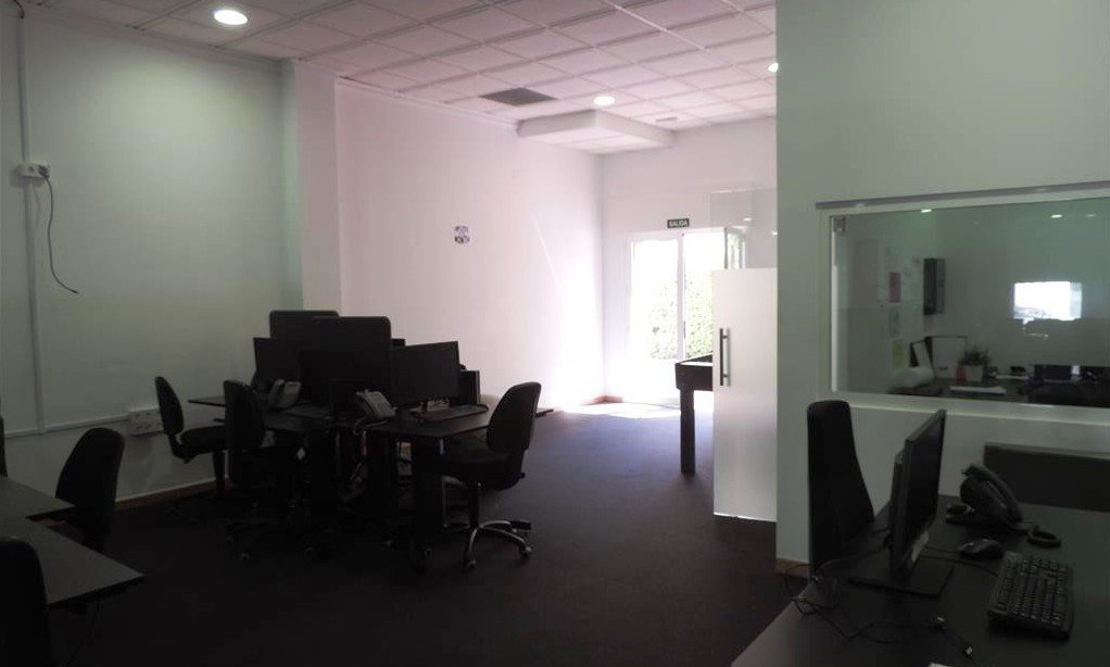 Modern office space with multiple computer workstations, chairs, and a glass-walled room visible.