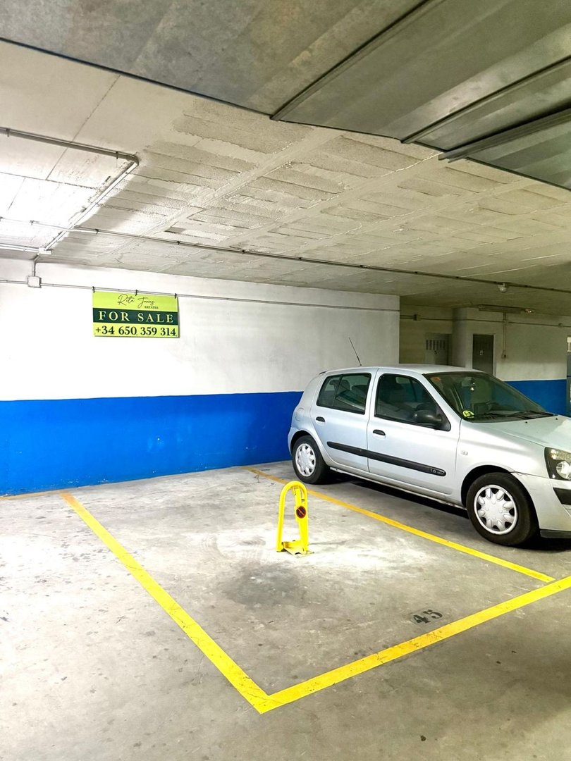 Underground parking space marked with yellow lines, featuring a silver car and a yellow bollard. Signage indicates 'For Sale'.