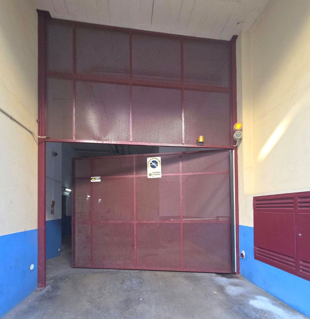 Metal industrial garage door with red mesh panels and warning signs, leading into a building.
