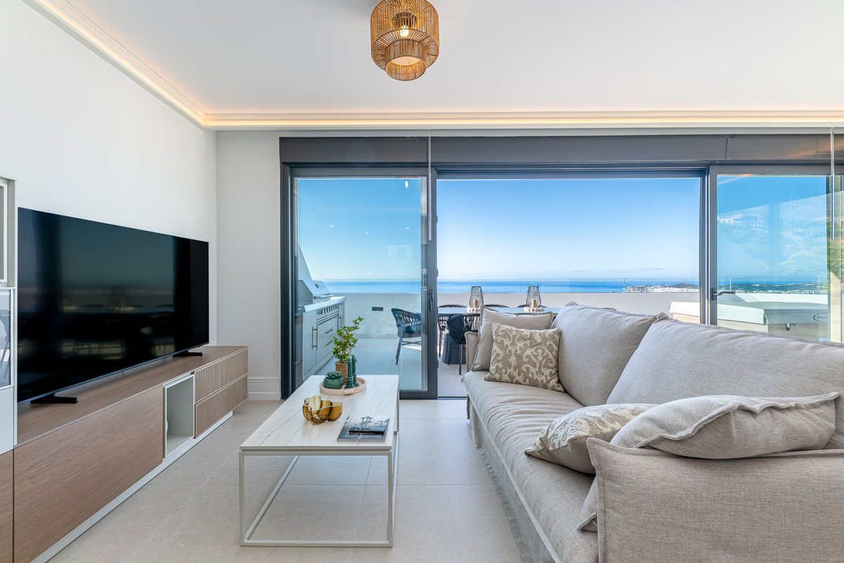 Bright living room with a sofa, coffee table, and a large sliding door opening to a balcony with an outdoor kitchen and sea views.