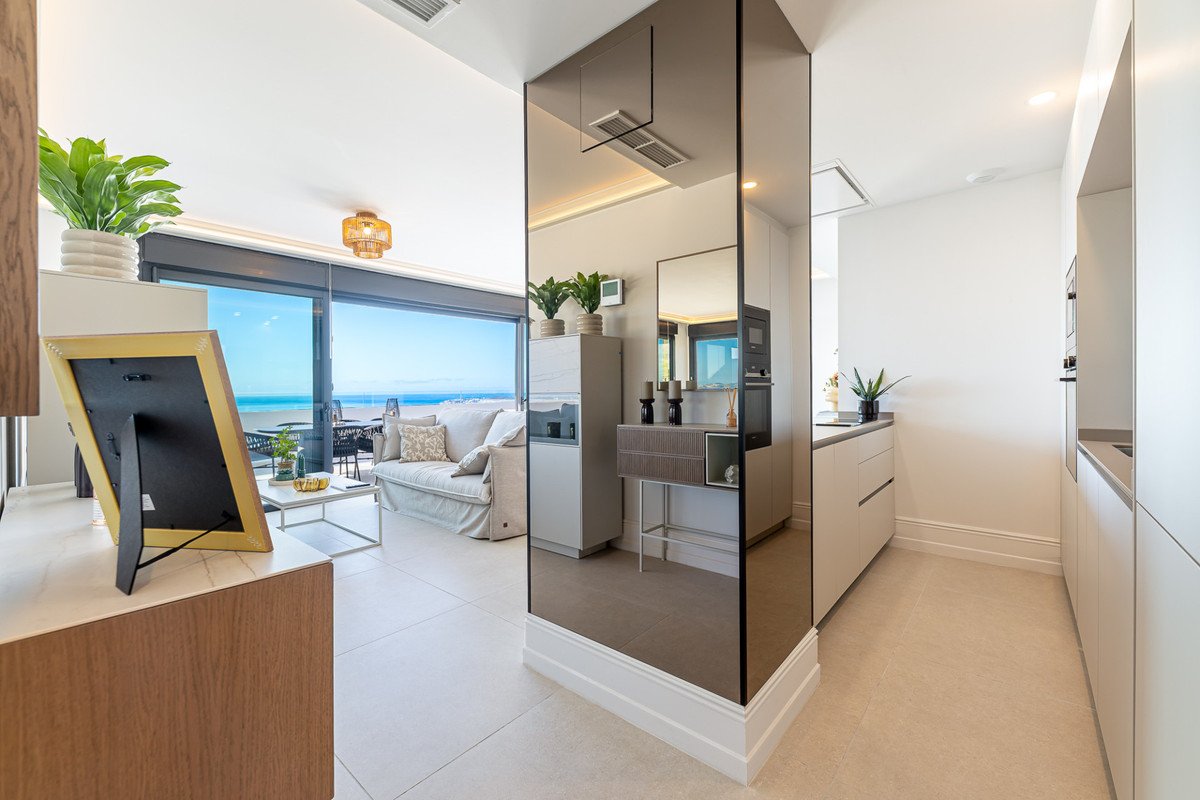 A modern living area with a sofa, coffee table, and a large window overlooking the sea. Includes a glimpse of a kitchen area.