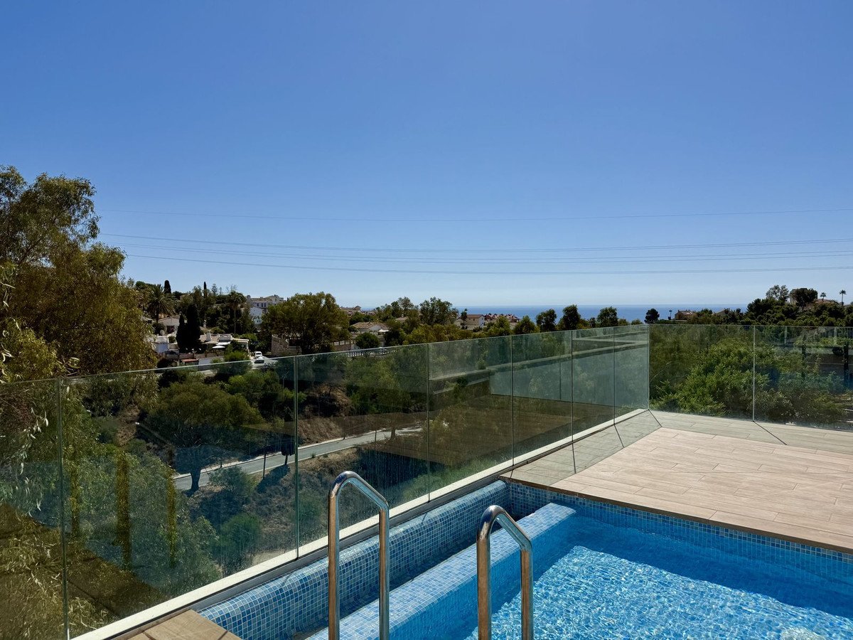 A modern private swimming pool with a wooden deck and glass railings, overlooking the sea and surrounding greenery.