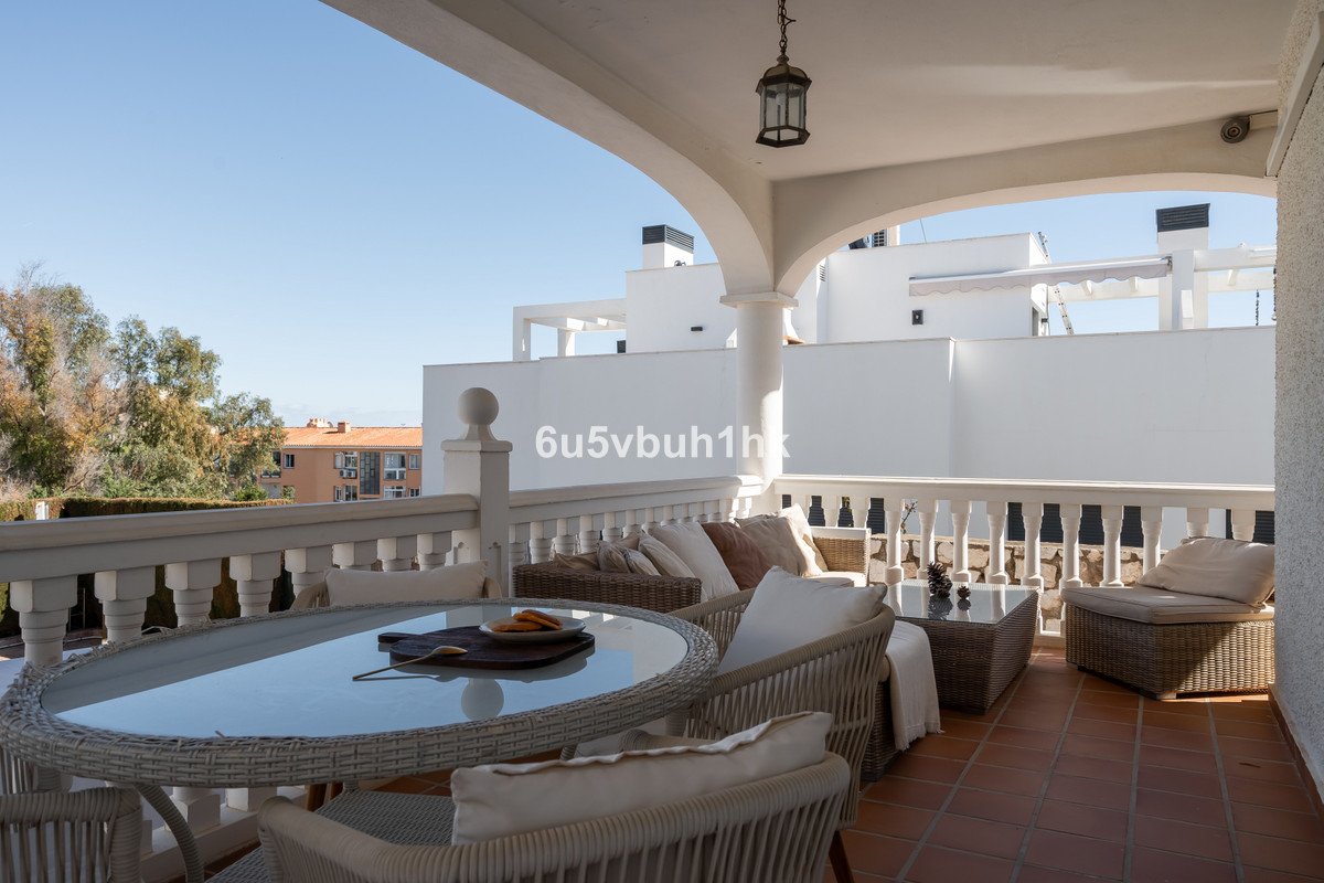 Covered balcony with wicker furniture, glass table, and white railings overlooking a sunny residential area.