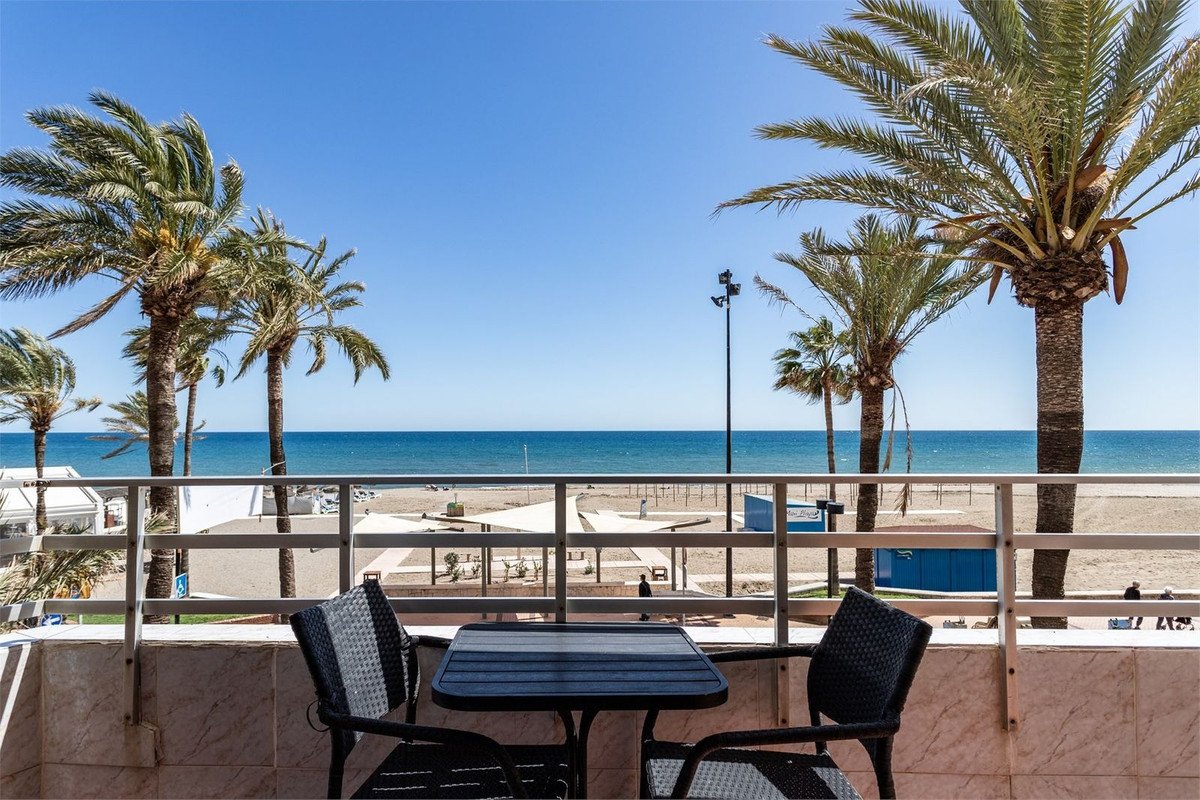 Balcony with a table and chairs overlooking the beach and sea with palm trees under a clear blue sky.