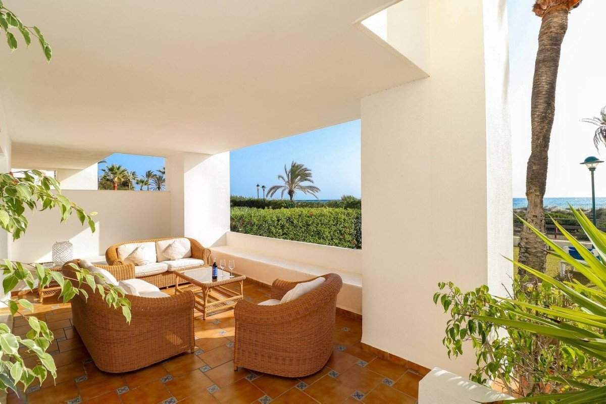 Terrace with wicker furniture, a table with wine, and a view of palm trees, green bushes, and the sea.