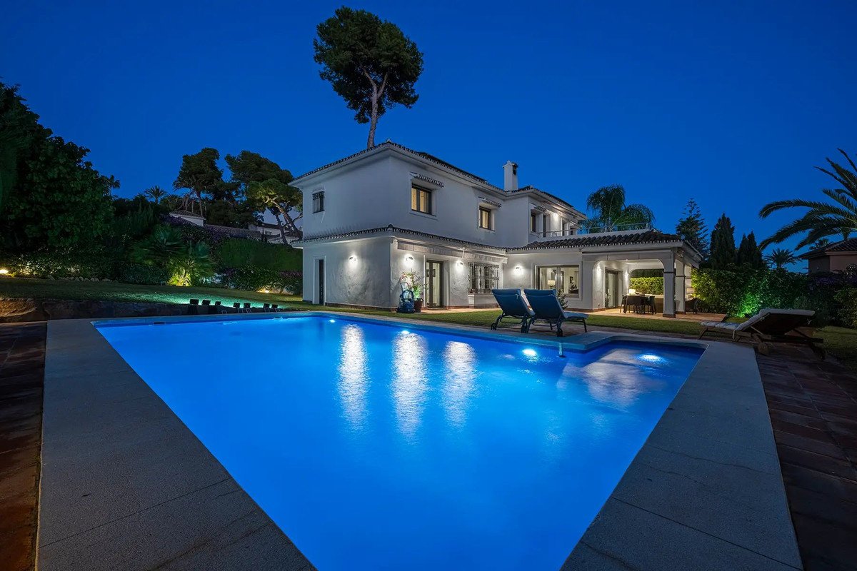 Luxury villa with a bright blue swimming pool and loungers, illuminated at dusk with garden lights.
