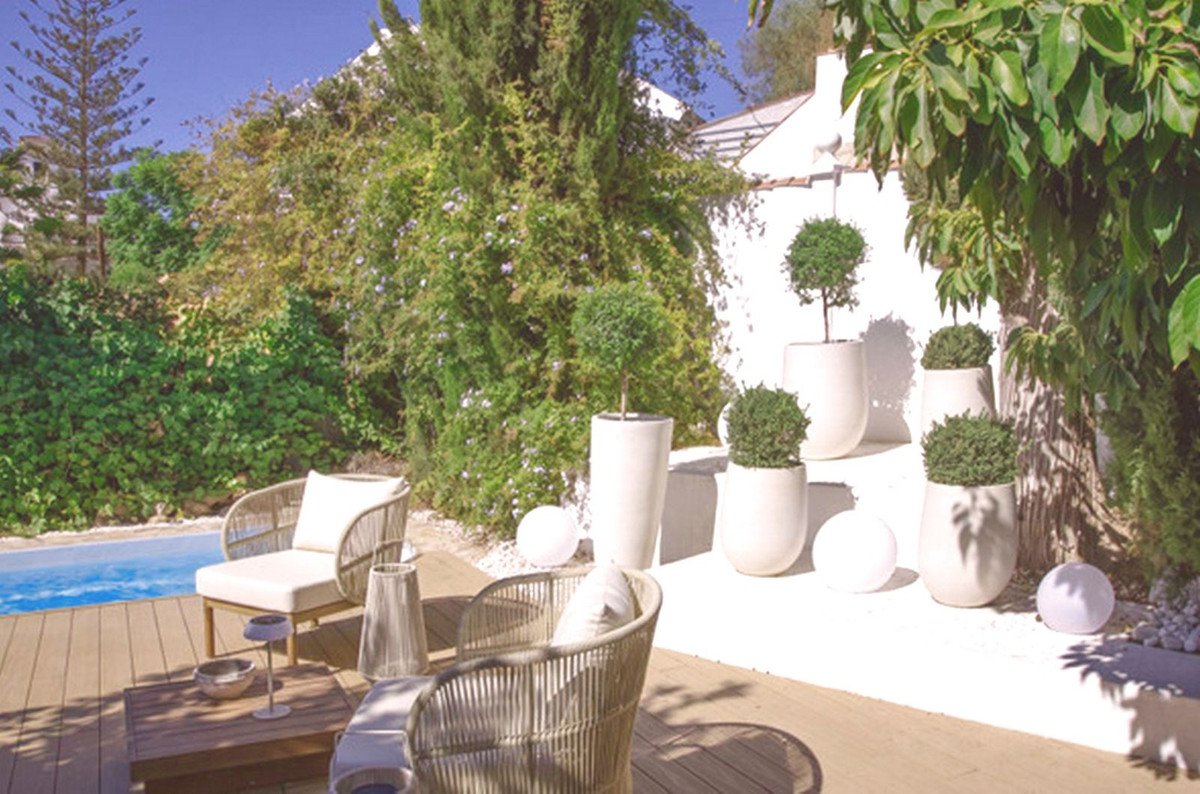 A patio with comfortable seating, decorative plants in pots, a swimming pool, and lush greenery.