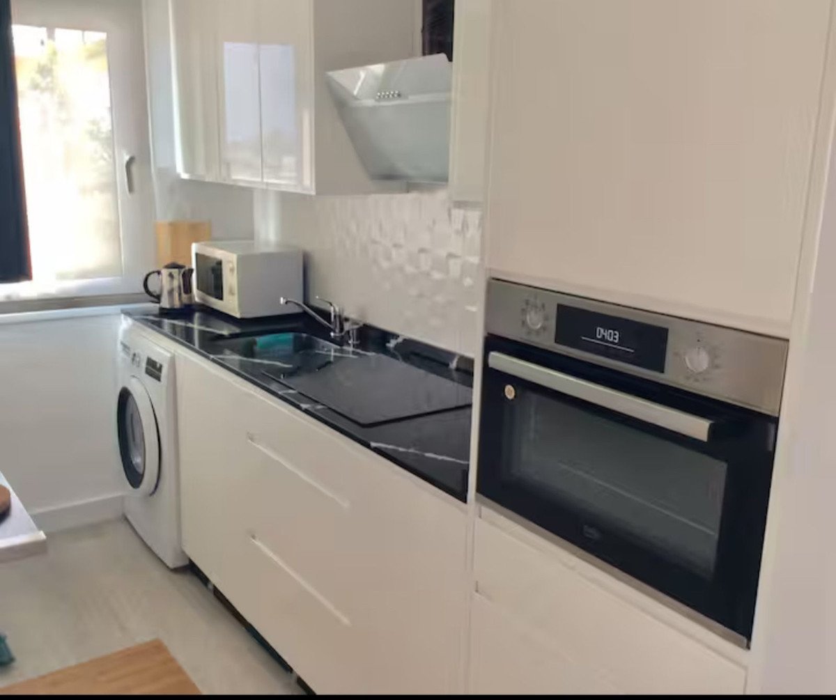 Modern white kitchen with black countertop, built-in oven, sink, microwave, and washing machine visible.