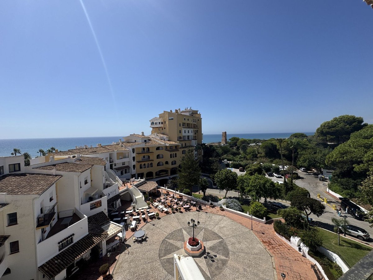 View from a balcony overlooking a courtyard with outdoor seating, buildings, trees, and the sea in the distance under a clear blue sky.