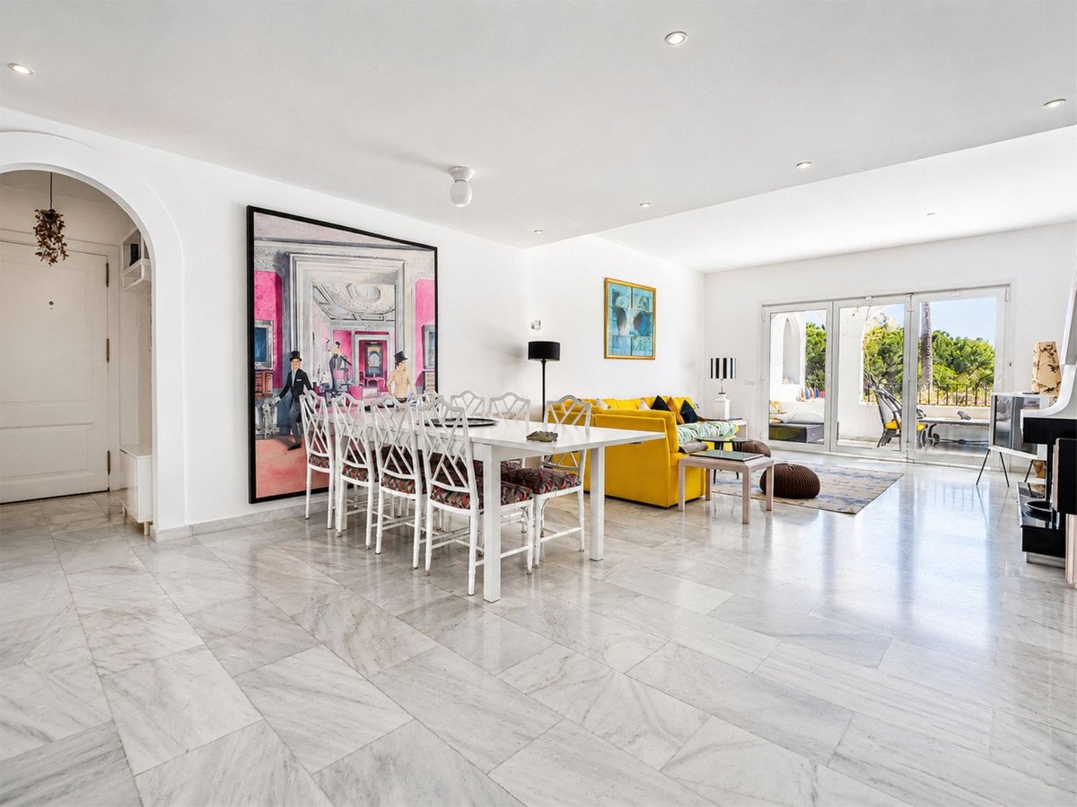 Spacious living and dining area with a yellow sofa, large abstract art, and patio doors leading to an outdoor space with a lush green view.