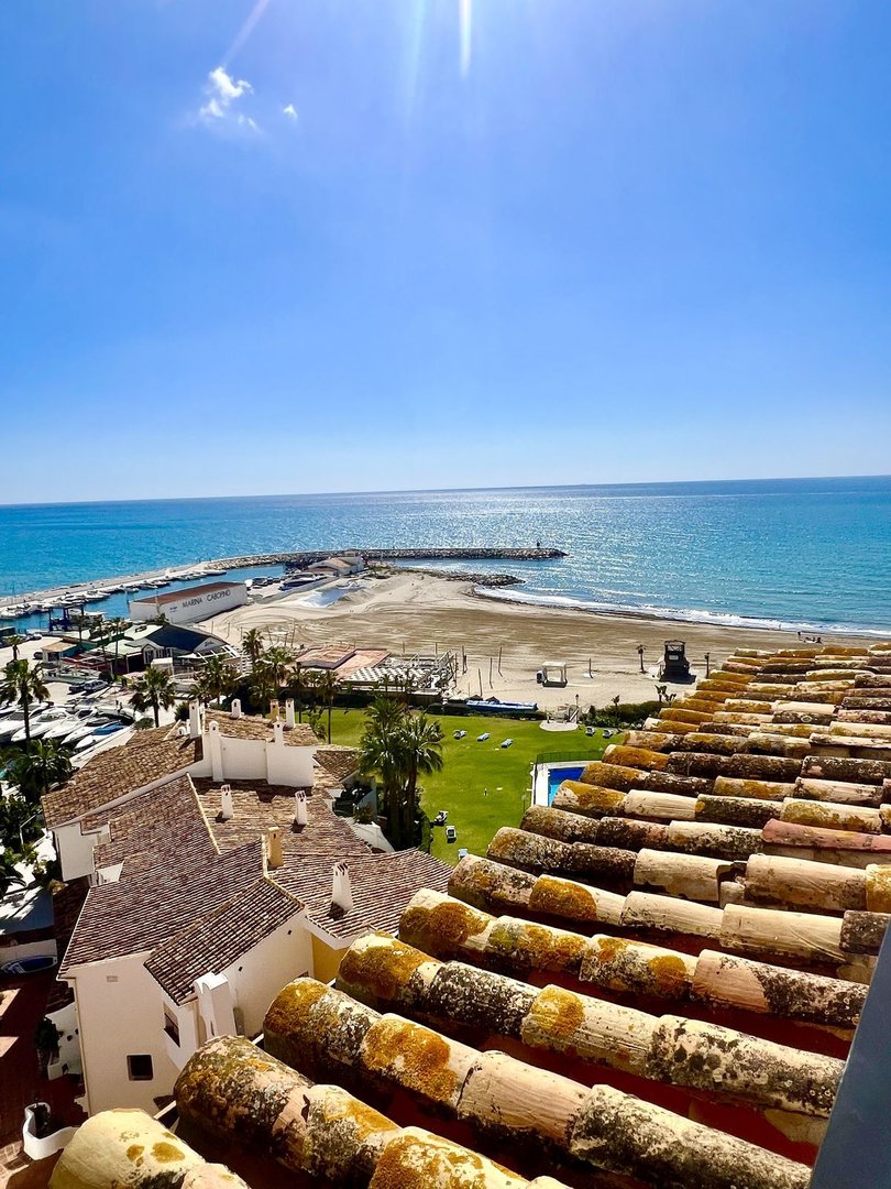 A sunny day with a view of the sea, beach, marina, and rooftops from a terrace.