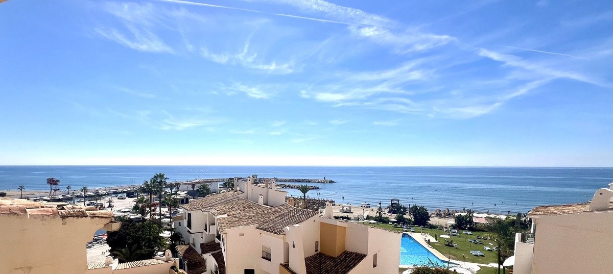 A scenic balcony view overlooking the sea, beach, and a swimming pool, with surrounding buildings and clear blue sky.