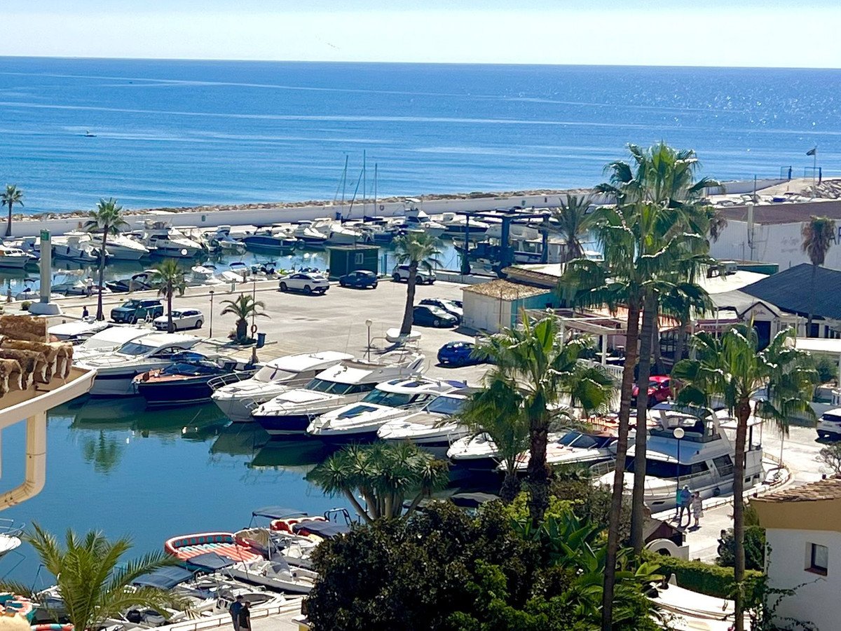 View of a marina with many boats docked, overlooking the calm blue sea and palm trees.