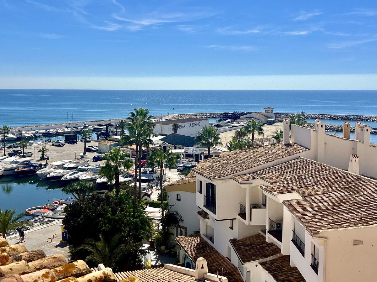 View from apartments overlooking a marina with boats, palm trees, and the sea under a clear blue sky.