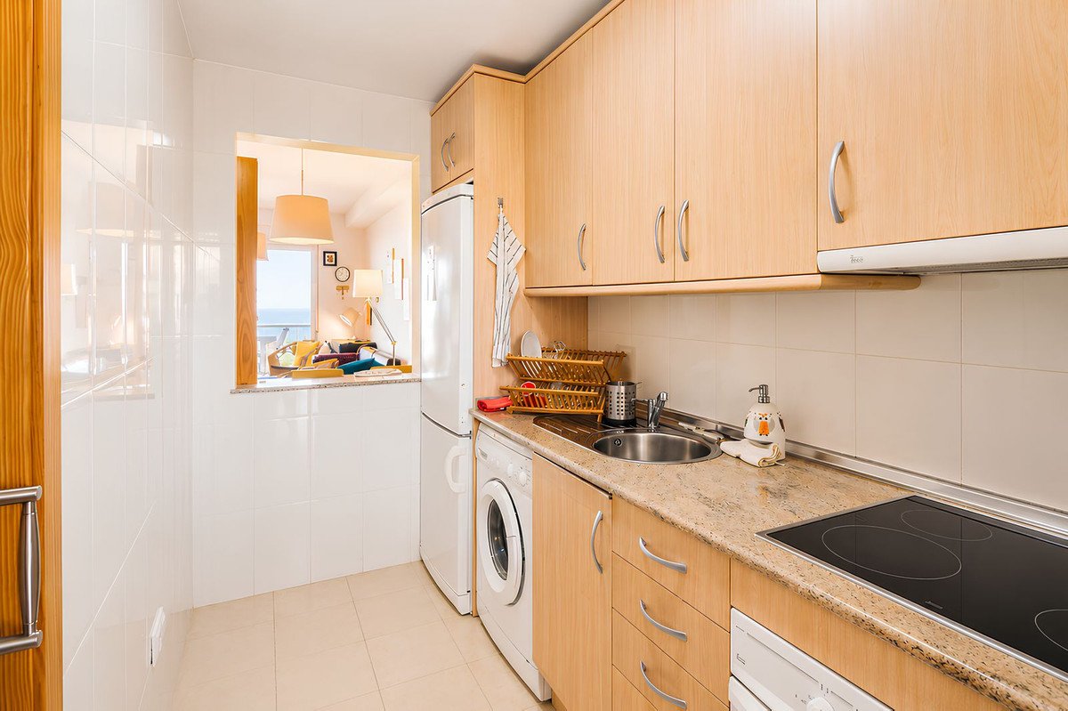Compact kitchen with light wood cabinets, a washing machine, and a window overlooking the sea and a living area.