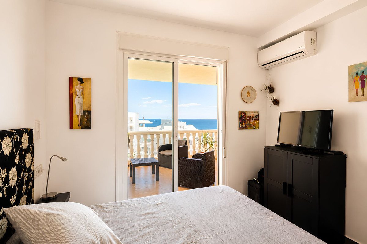 Bedroom with a bed, TV, and air conditioning, opening to a balcony with chairs and a sea view.