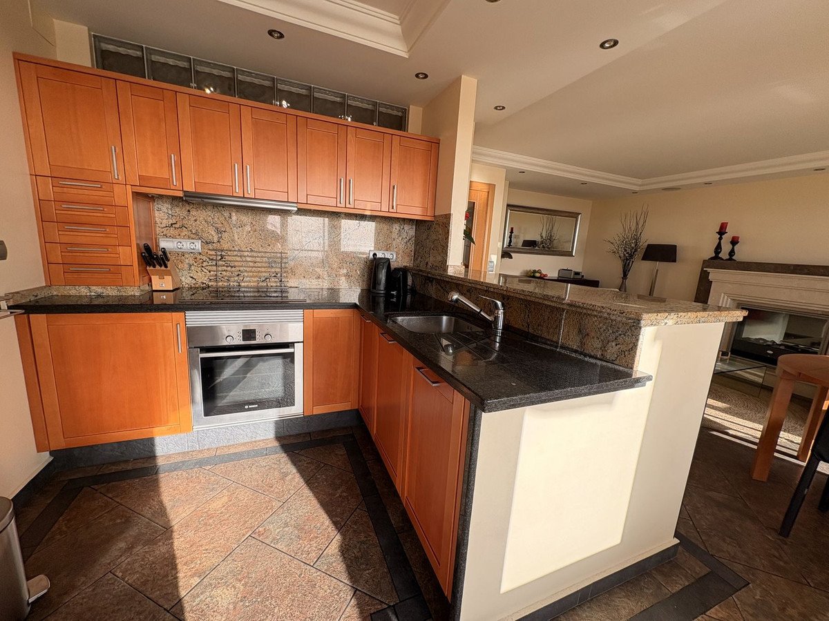 A modern kitchen with granite countertops, sleek wooden cabinets, and an integrated oven. integrated oven. oven. Adjacent living area with fireplace visible.