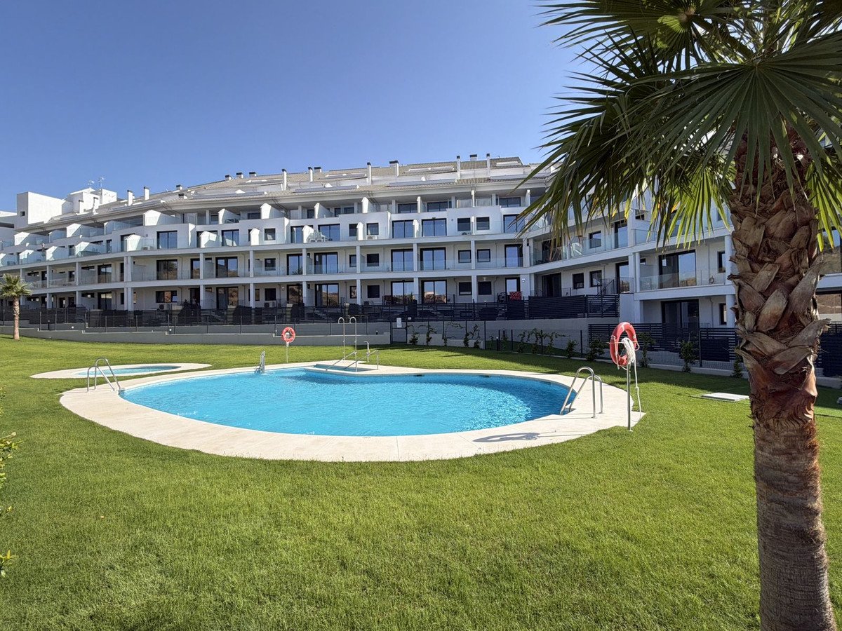 A modern apartment complex with balconies and a swimming pool in front, featuring a palm tree on the right and a clear blue sky above.