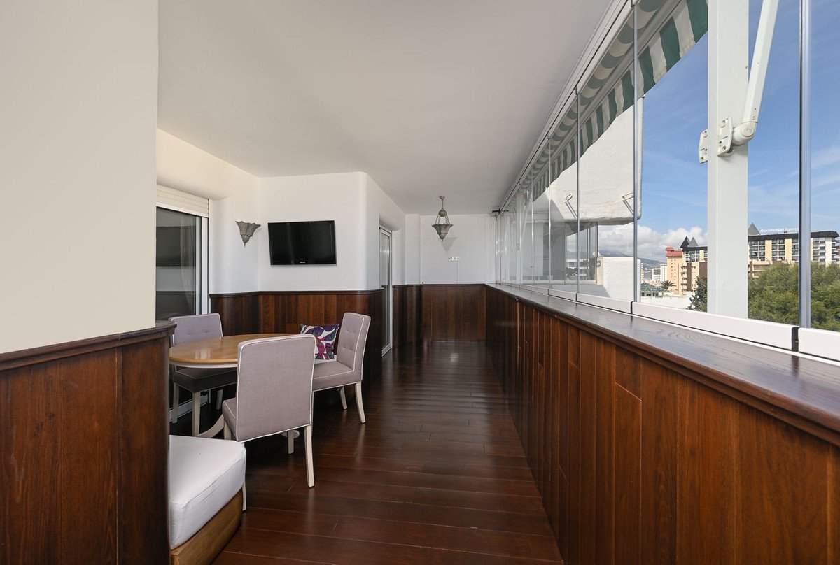Long balcony with wooden flooring, glass railing, and city view, featuring a dining table and TV.