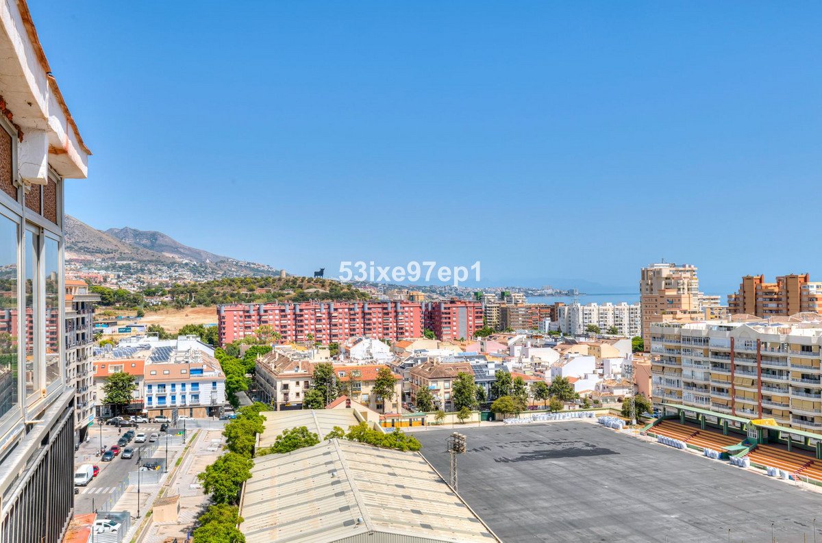 View from a balcony overlooking a city with mountains and the sea in the distance, under a clear blue sky.