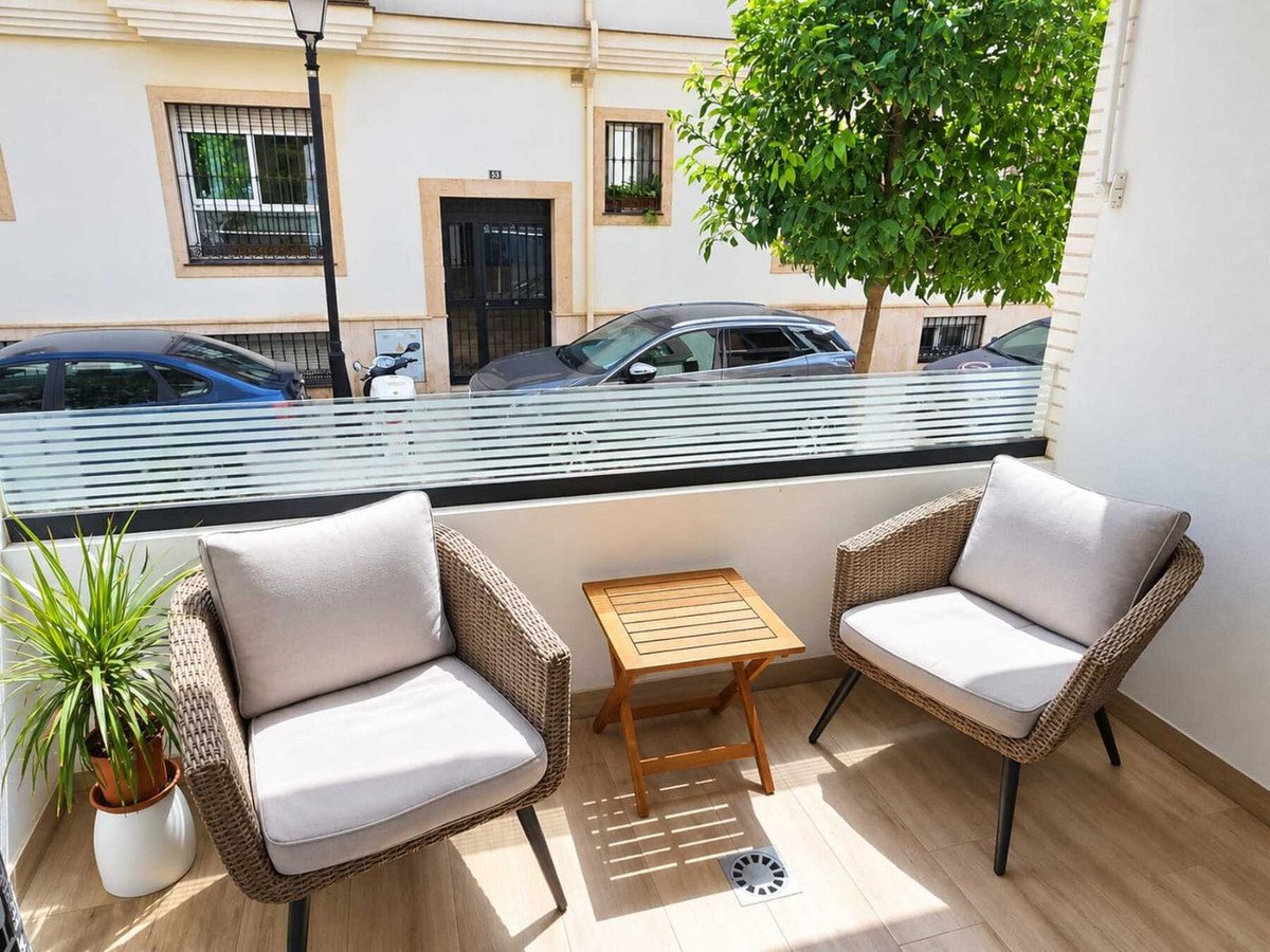 A cozy balcony with two wicker chairs and a small wooden table, overlooking a street with parked cars and a tree.
