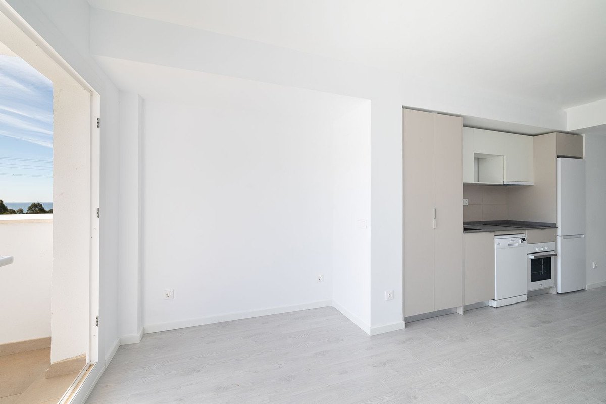 A modern studio apartment with a kitchenette, balcony, and a view of the sea and distant trees.